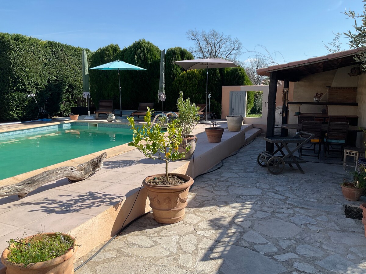 The outdoor area features a swimming pool surrounded by well-maintained hedges. Sun umbrellas provide shade over seating areas, while potted plants enhance the space. A covered area with seating and a stone path leads towards the pool, creating a tranquil environment for relaxation.