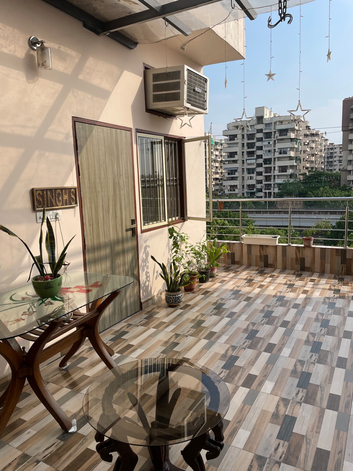 An outdoor balcony features a glass table and wooden chairs. Potted plants are arranged along the railing. The flooring displays a modern tile pattern, and a window with metal grilles allows for natural light. Nearby buildings can be seen in the background.
