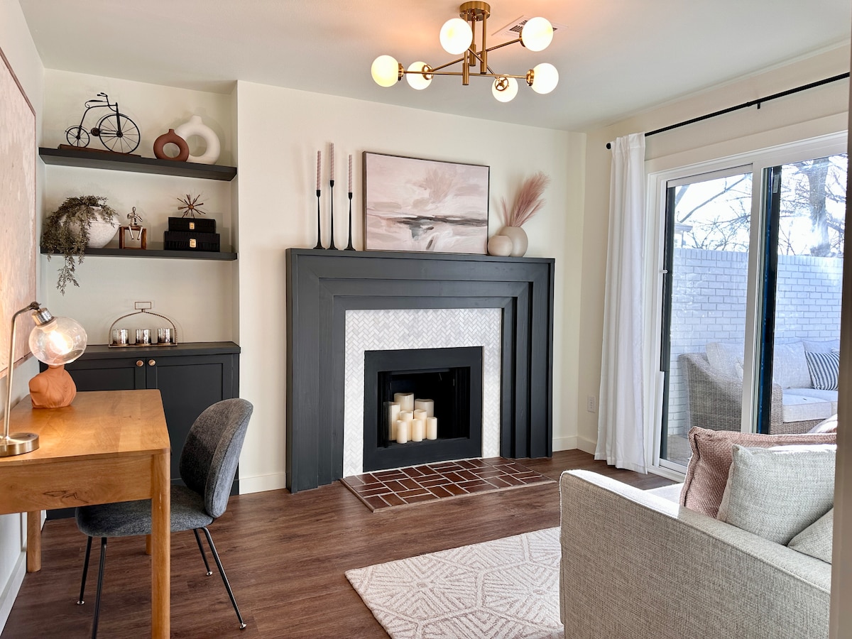 A modern living area features a decorative fireplace with candles nestled inside. A desk and chair are positioned nearby, while a large window with sheer curtains allows natural light to fill the room. Soft furnishings and neutral colors create a calming environment.