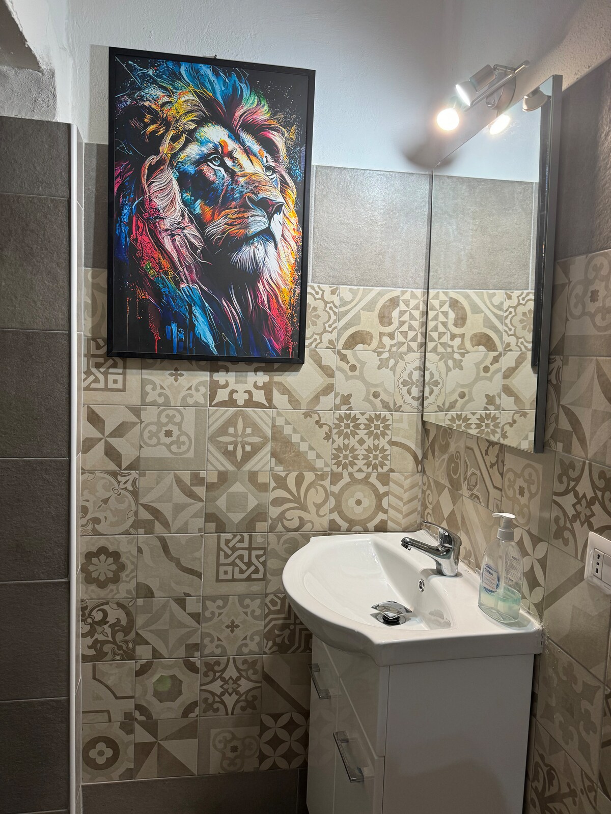The bathroom showcases a modern sink with a clean white finish beneath a colorful lion artwork. The walls are adorned with a mix of patterned tiles in earthy tones, creating a vibrant yet balanced atmosphere. A well-placed light fixture provides illumination.