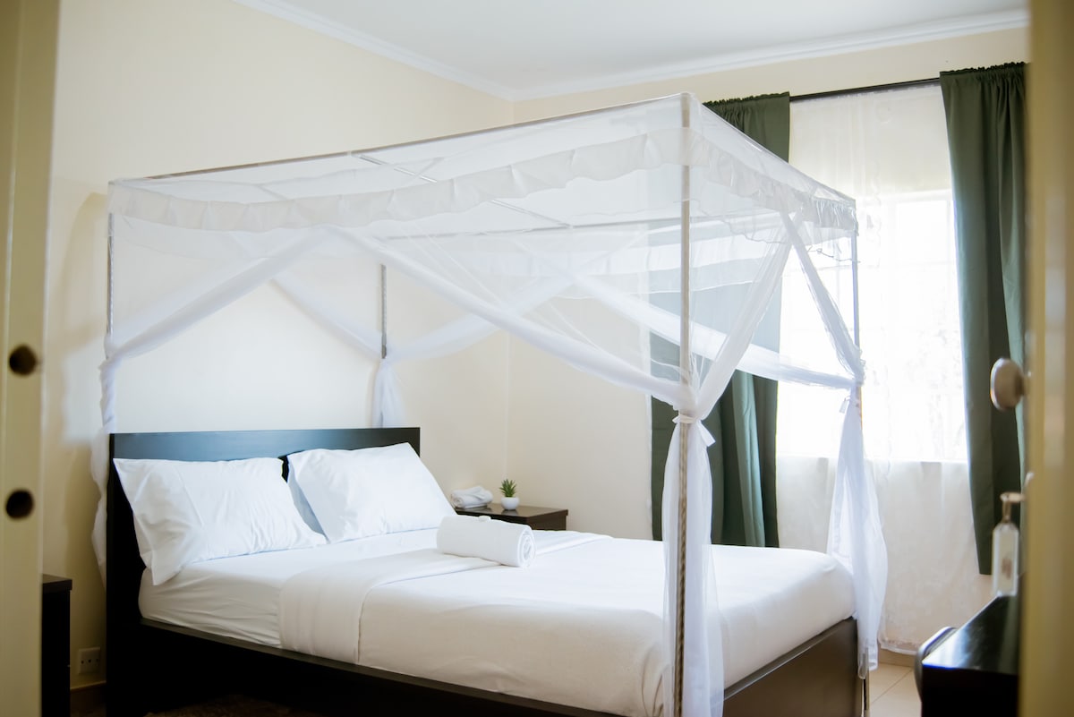 A comfortable room features a queen bed framed by mosquito netting. The bed is complemented by a towel neatly rolled on the bedding. Large windows adorned with dark curtains allow natural light to filter in, enhancing the serene atmosphere.