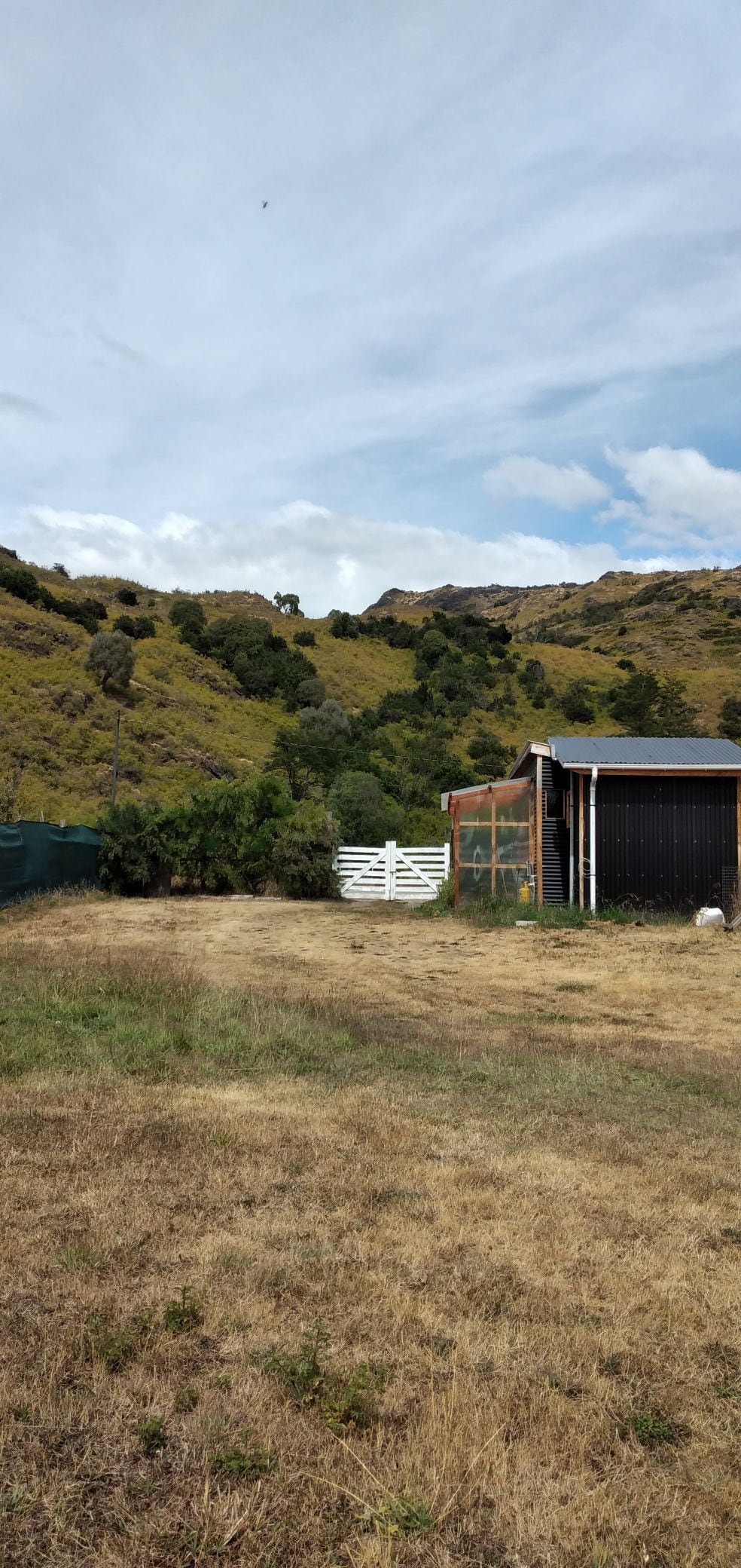 A rustic property is outlined by natural landscapes featuring rolling hills and vegetation. A white gate stands prominently in view, leading to a grassy area, while a simple structure accompanies the land, suggesting a serene and secluded environment.