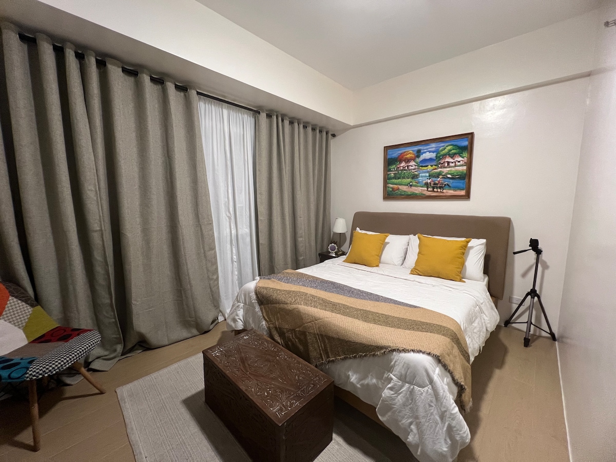 A brightly lit bedroom features a spacious bed with neutral-toned bedding and two yellow accent pillows. Large curtains frame the window, while a colorful patterned chair and a decorative wooden trunk add interest to the space. A framed landscape artwork is displayed on the wall.