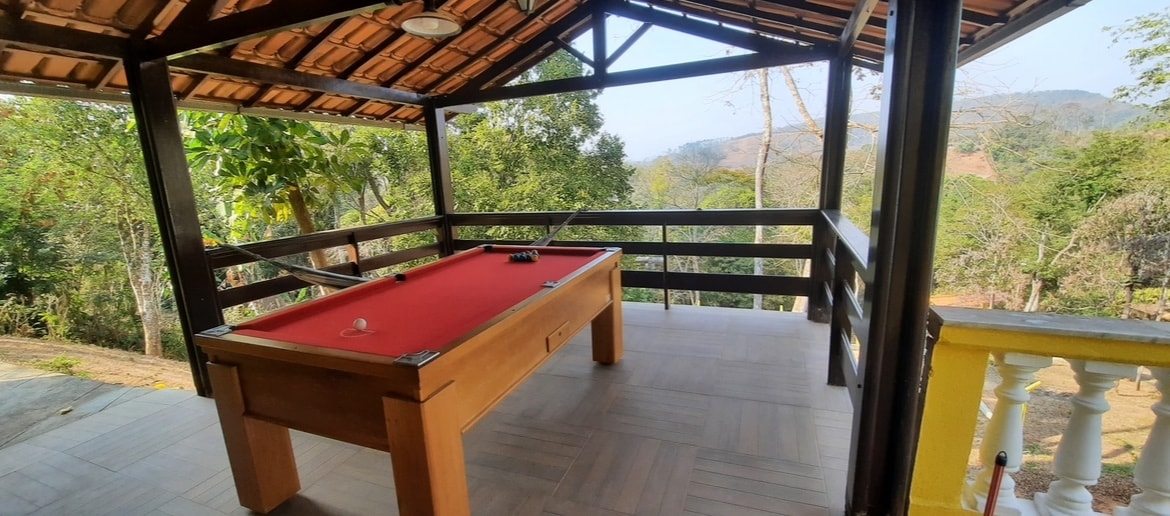 An outdoor game area features a well-maintained pool table under a wooden roof. Surrounding trees provide a natural backdrop, while expansive views of distant hills are visible through the open sides, creating a pleasant space for relaxation and entertainment.