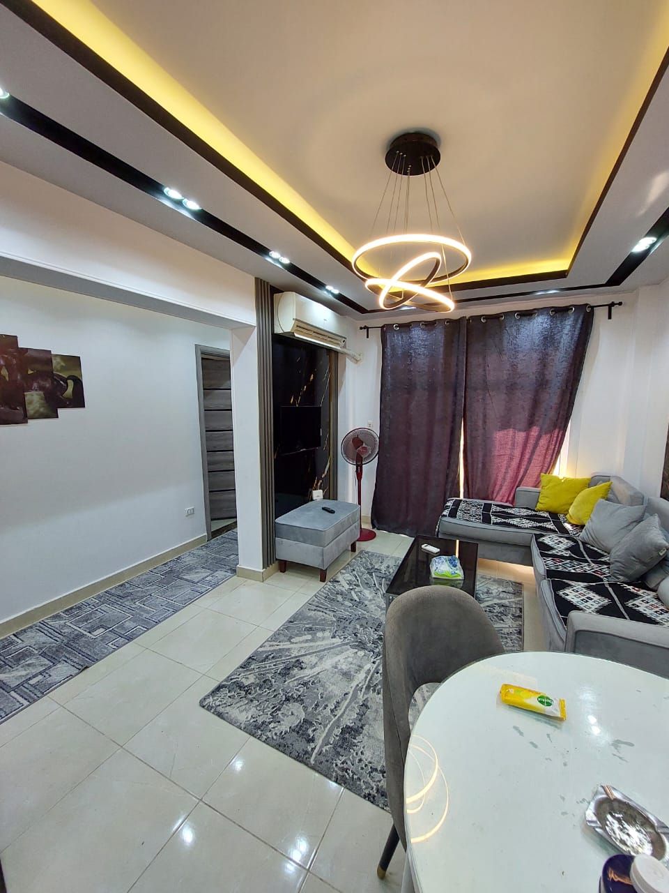 An elegant living space features a modern seating area with a plush gray sofa and decorative pillows. A dining table is set for four. Natural light enters through dark, sheer curtains, and modern lighting fixtures enhance the atmosphere. A patterned rug adds warmth to the tiled floor.