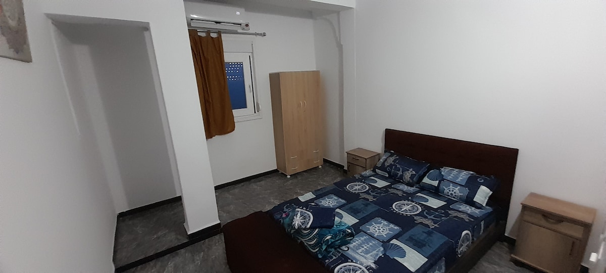 A spacious bedroom is presented with a large bed featuring a blue patterned comforter. Two bedside tables are positioned on either side, and a wardrobe stands nearby. A window with light drapes allows natural light into the room.