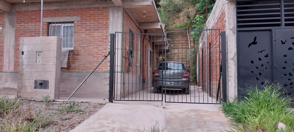 A gated entrance reveals a car parked in a driveway, flanked by low green grass. The brick facade of the house is visible on one side, with an adjacent wall displaying a modern design. The surrounding area is slightly elevated, leading to a hillside.