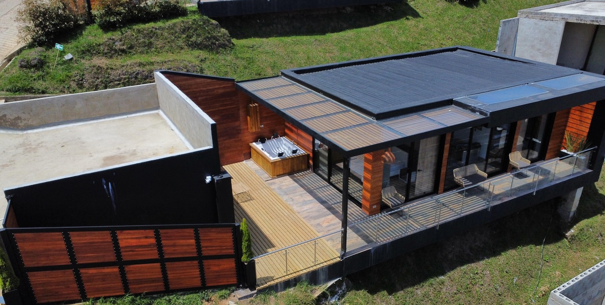 A contemporary single-story home features a flat roof and large glass sliding doors that provide access to an expansive deck. The outdoor area includes a built-in grill, while wooden slats add texture to the design, enhancing the overall modern aesthetic.