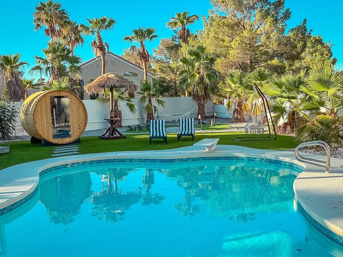 A sparkling pool reflects the vibrant blue sky, surrounded by lush tropical palm trees. Comfortable lounge chairs are positioned by the edge, and a unique wooden sauna can be seen in the background, adding to the serene outdoor atmosphere of the space.