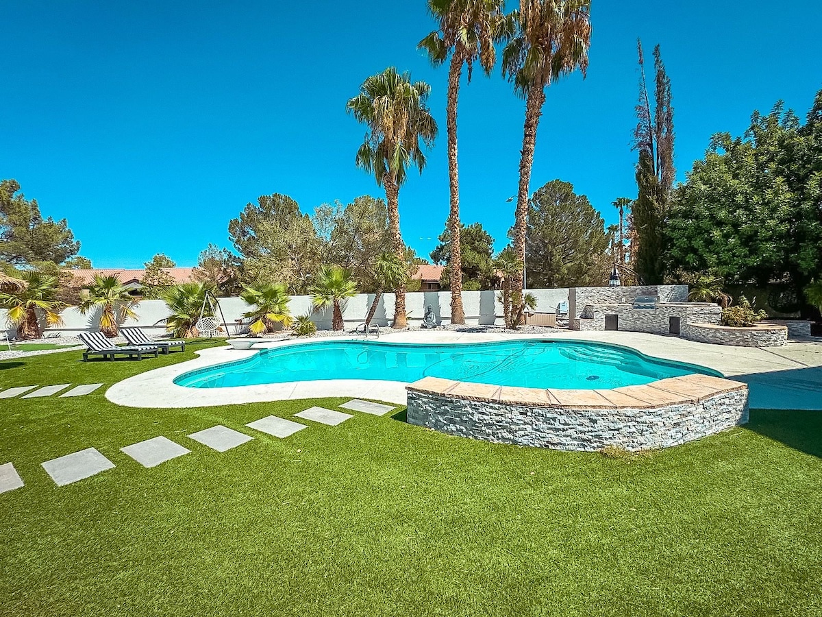 A large, inviting swimming pool is surrounded by a beautifully manicured grassy area. Tall palm trees provide shade while lounge chairs are positioned nearby for relaxation. Stone pathways lead around the pool, enhancing the serene outdoor atmosphere.