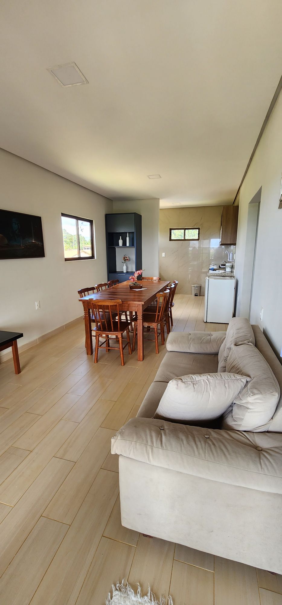 An open living and dining area is displayed, featuring a wooden dining table surrounded by chairs. Light-colored walls are complemented by a comfortable sofa nearby. A kitchen area can be seen in the background, with natural light entering through large windows.