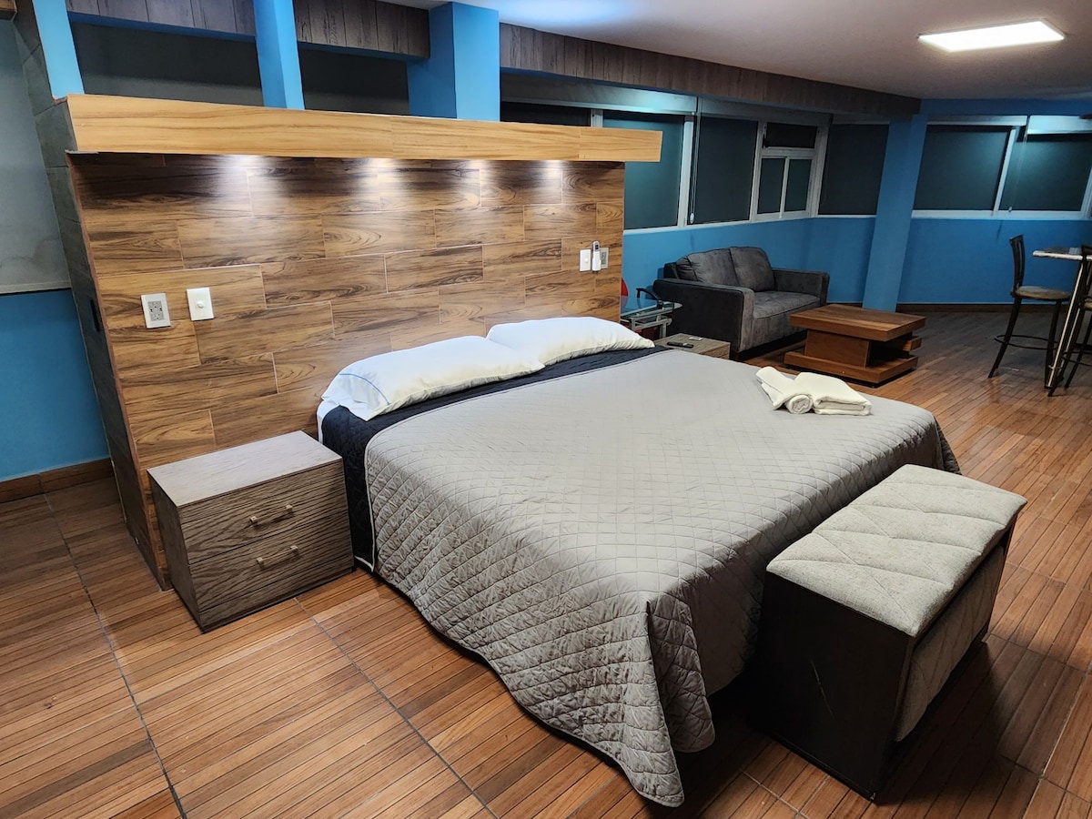 A spacious sleeping area features a large bed with a textured gray blanket and a wooden headboard. A comfortable bench sits at the foot of the bed, and natural light enters through the nearby windows. A seating area with a sofa and coffee table is visible in the background.