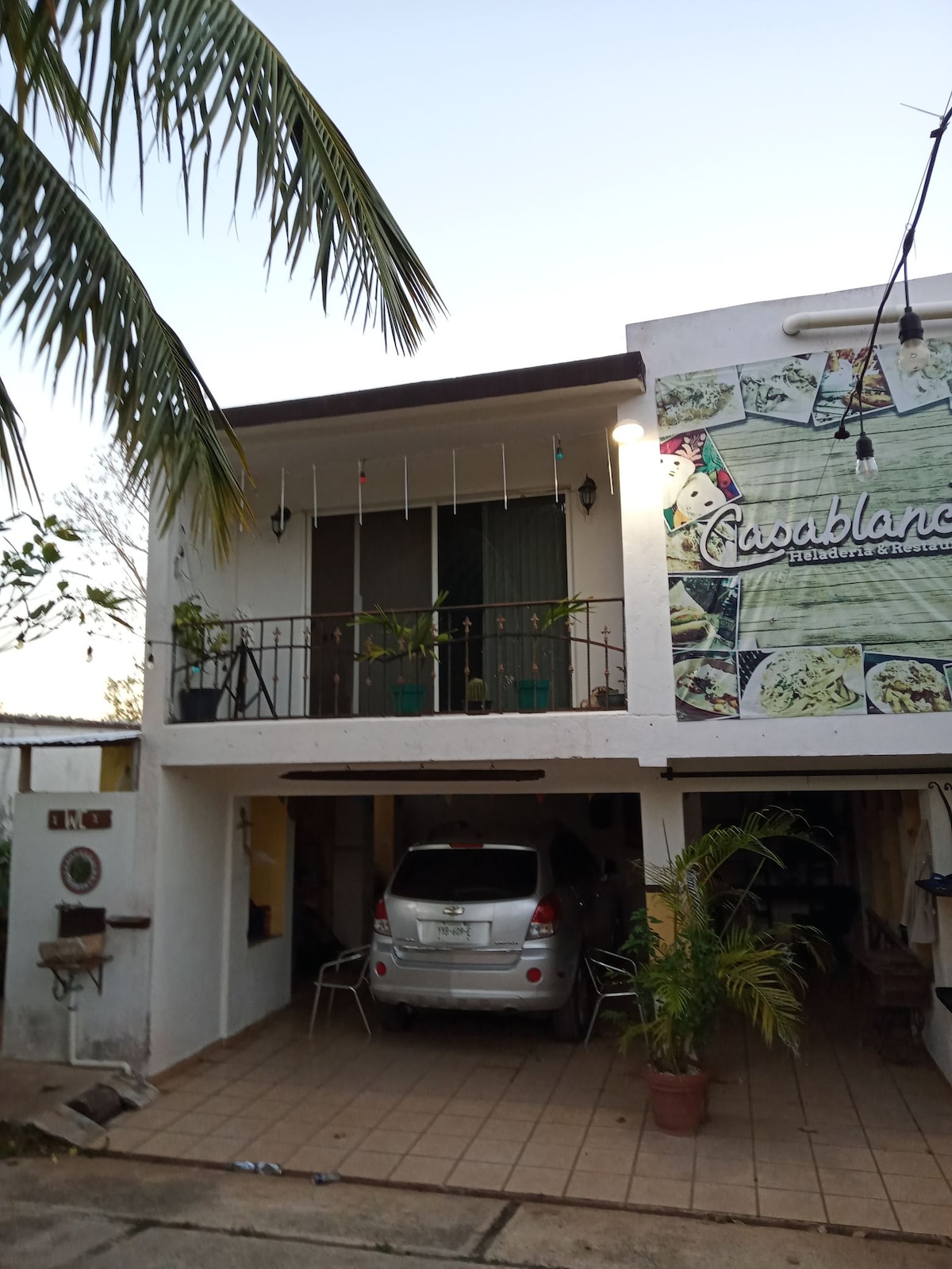 A two-story building features a balcony with decorative green drapes and potted plants. The lower level includes a tiled outdoor area, with a parked vehicle and metal chairs visible. The exterior is adorned with vibrant imagery related to local cuisine.