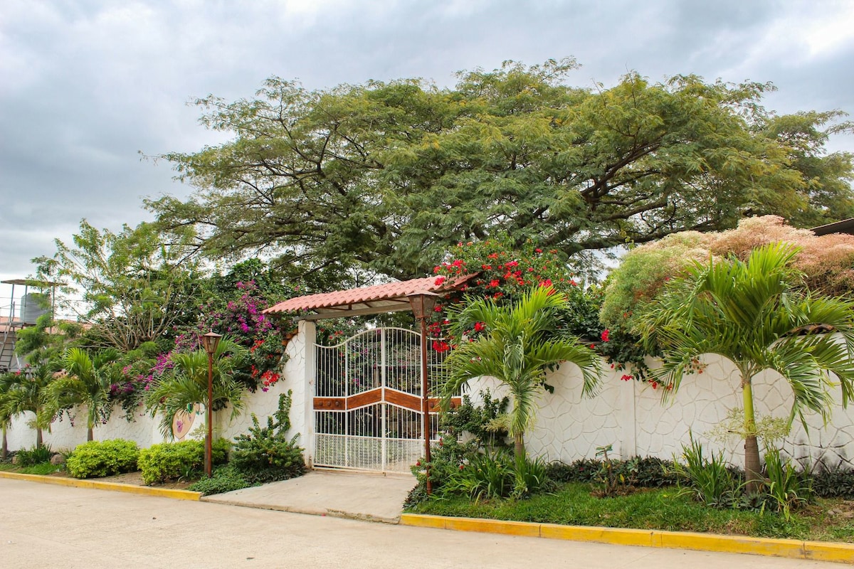 A gated entrance is surrounded by lush greenery and vibrant flowering plants. A decorative white wall with a textured design enhances the privacy of the property. Overhead, a large tree offers shade, contributing to a serene outdoor atmosphere.