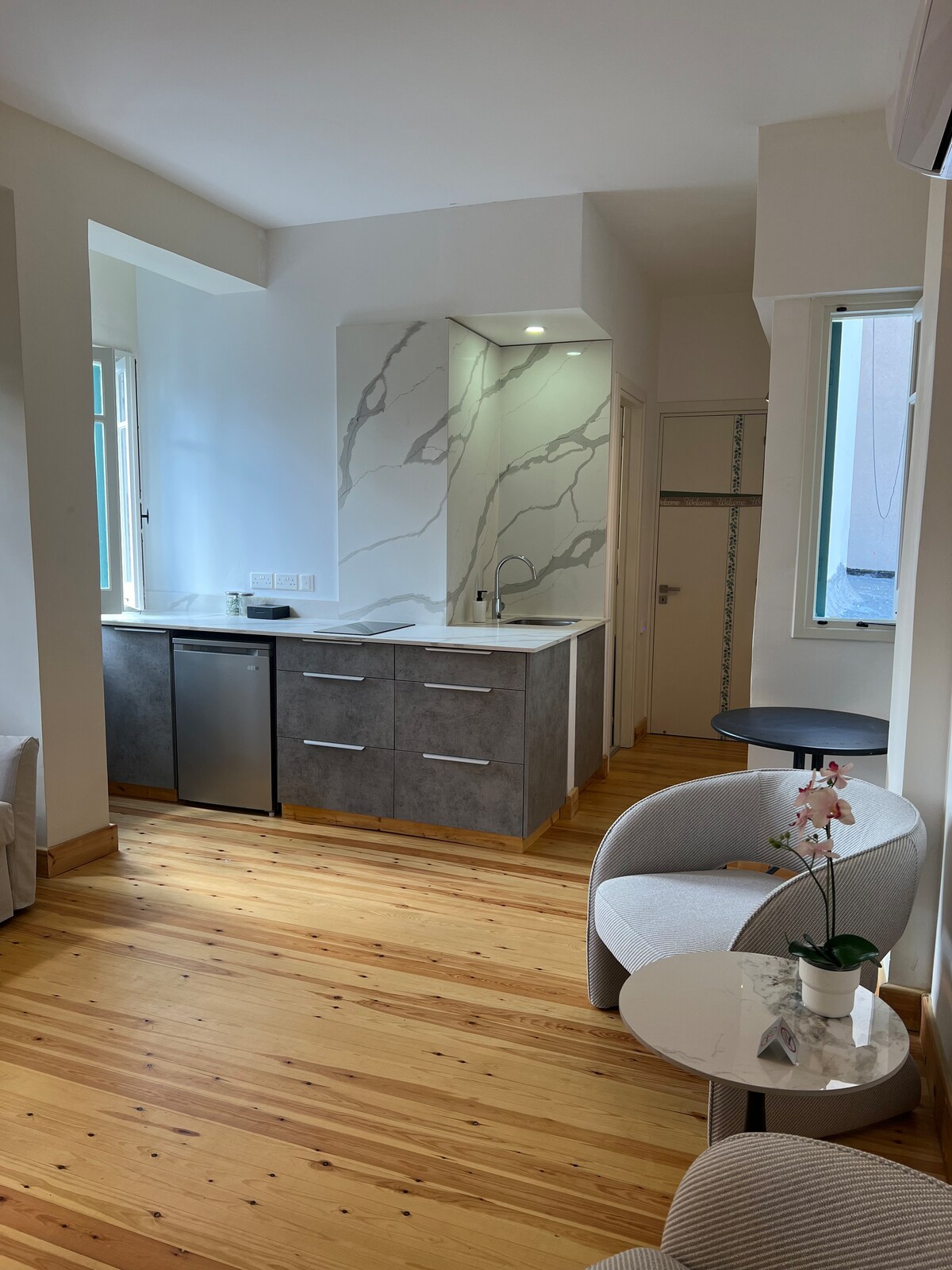 A modern kitchen area features sleek cabinetry, a stainless steel refrigerator, and a marble backsplash. Natural light filters through large windows, illuminating the light wooden flooring. A small round table and comfortable seating create a cozy atmosphere in the open space.