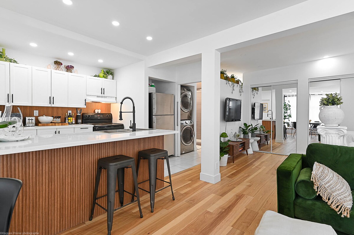 The open concept space features a modern kitchen with a large island, two black bar stools, and ample storage. Natural wood accents complement the sleek white cabinetry. A cozy green sofa is positioned nearby, while sunlight brightens the room through large windows.