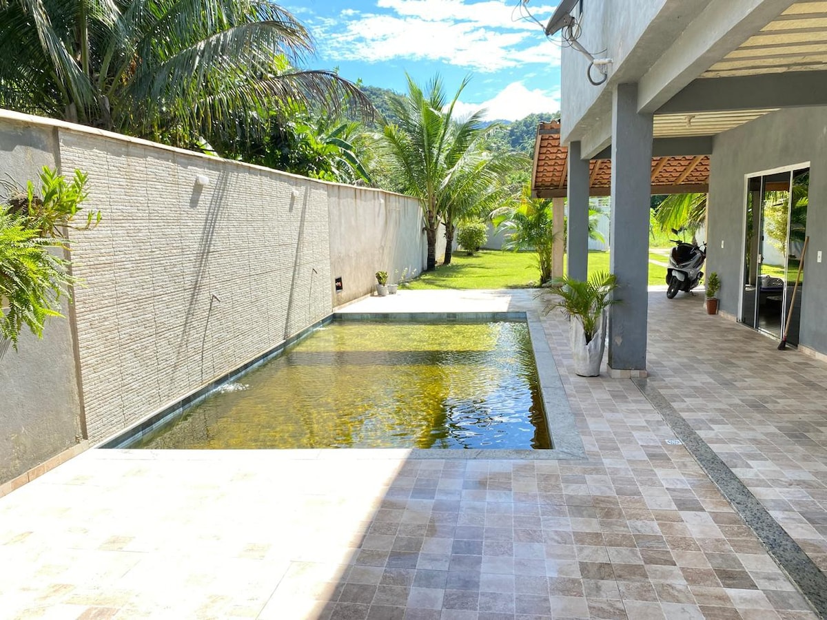 A serene outdoor area features a small pool surrounded by vibrant greenery. The stone patio is complemented by a smooth walkway that connects the pool to the adjacent covered space, offering a view of the garden and nearby palm trees.