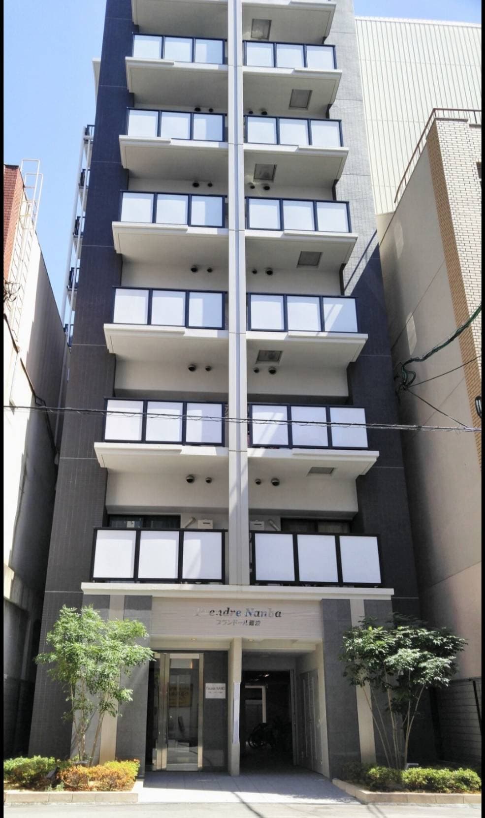 A modern building with a grey exterior features multiple floors and large windows providing ample natural light. The entrance is framed by small trees, adding a touch of greenery to the urban setting. The façade presents a clean and organized appearance.