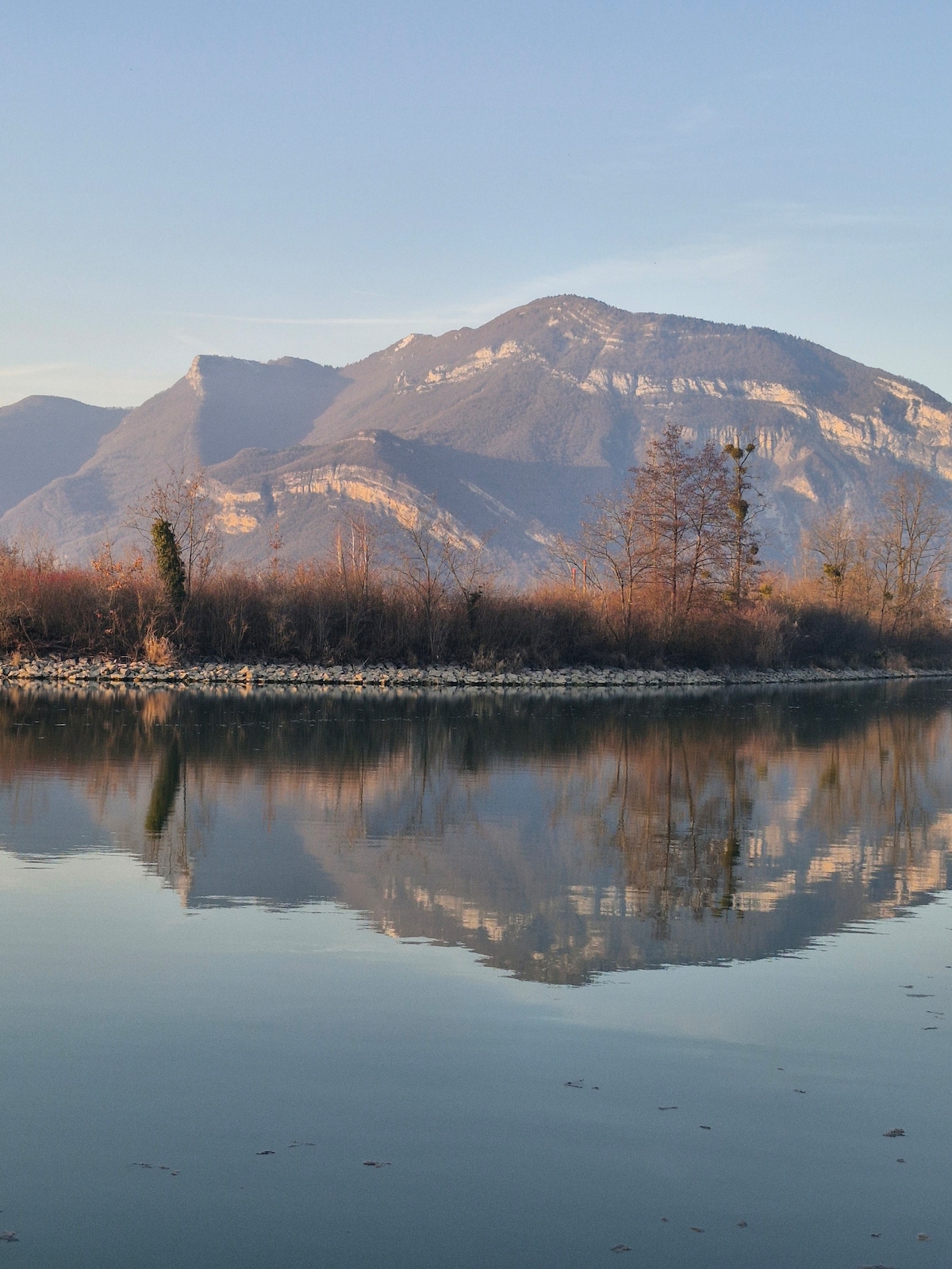 A serene landscape features a mountain range reflected in calm waters. The mountains display varying shades of brown and green, while the water's surface creates a mirror-like effect. Sparse vegetation lines the shore, enhancing the natural tranquility of the scene.