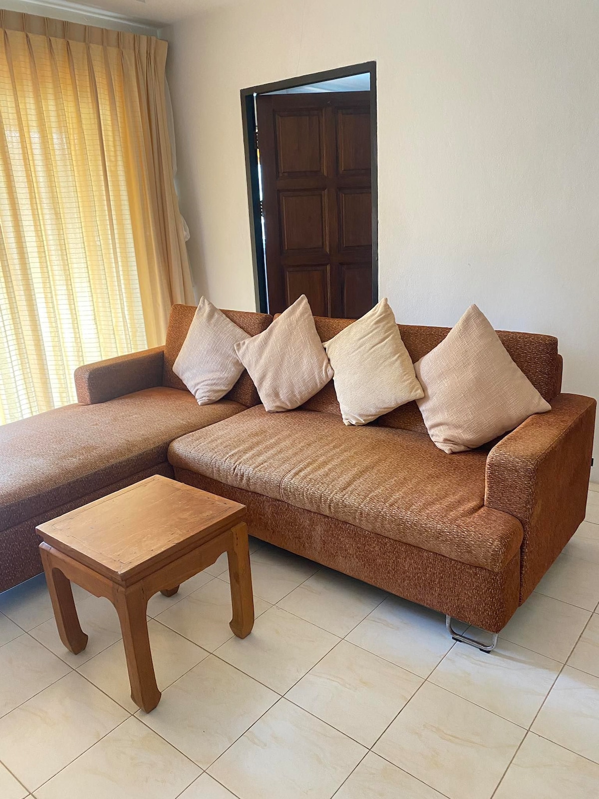 A comfortable living area features a brown sectional sofa adorned with three light-colored throw pillows. A small wooden side table stands nearby. Soft natural light enters through the window, which is lined with light yellow curtains, creating an inviting ambiance.