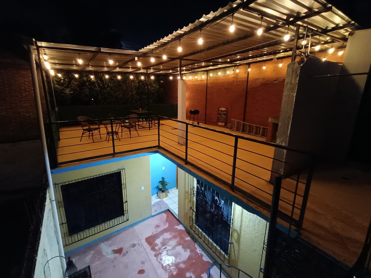 A spacious outdoor terrace features string lights overhead, creating a warm ambiance. Several chairs and a small table are arranged for outdoor dining. The walls are painted in vibrant colors, and a section of the lower patio is visible, enhancing the inviting atmosphere.