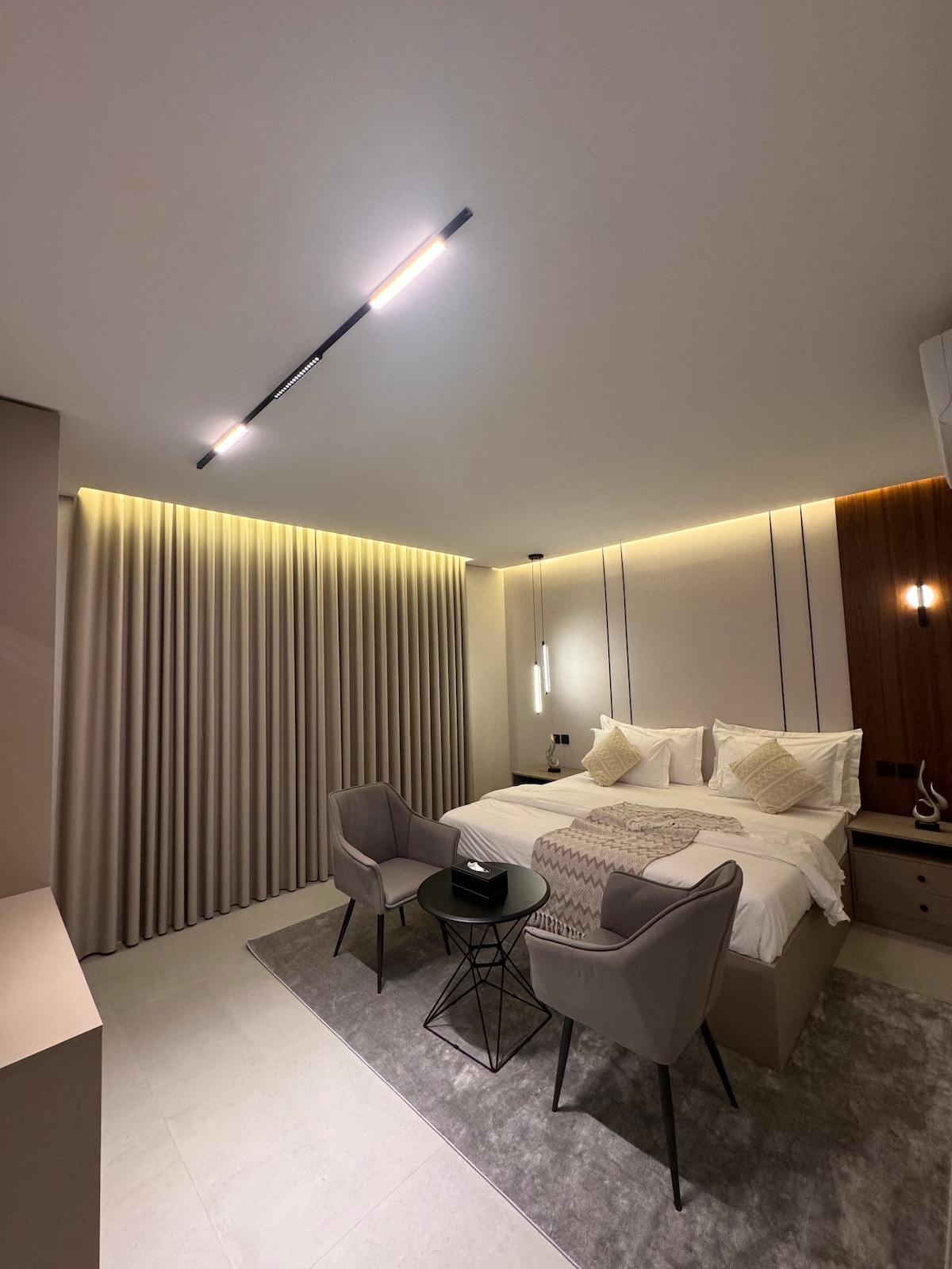 The bedroom features a spacious bed with soft linens, flanked by contemporary bedside tables. Two stylish chairs and a small table are set on a plush rug, while warm indirect lighting highlights the textured walls and floor-to-ceiling curtains, creating a calming ambiance.