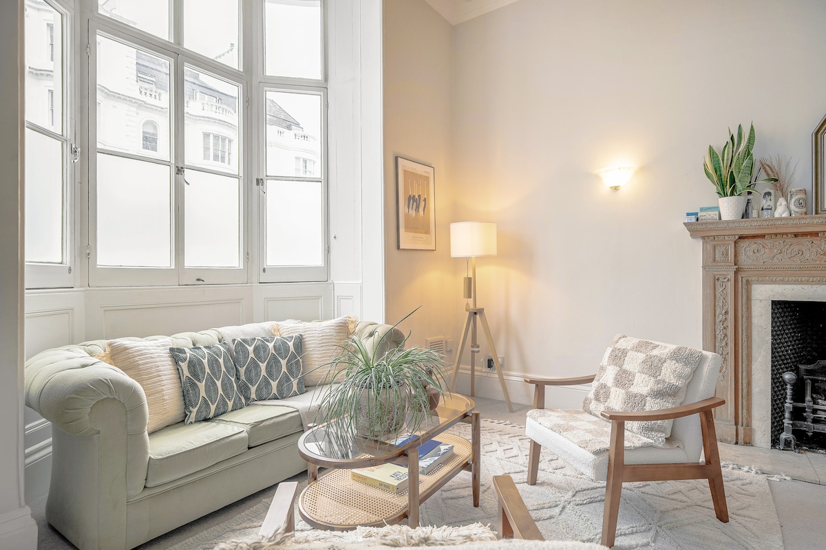 A bright living area features a light-colored sofa adorned with decorative cushions, complemented by a wooden armchair. A glass coffee table holds books and a plant. Natural light streams through tall windows, illuminating the space. Decorative elements include a lamp and a fireplace surrounded by a detailed mantel.