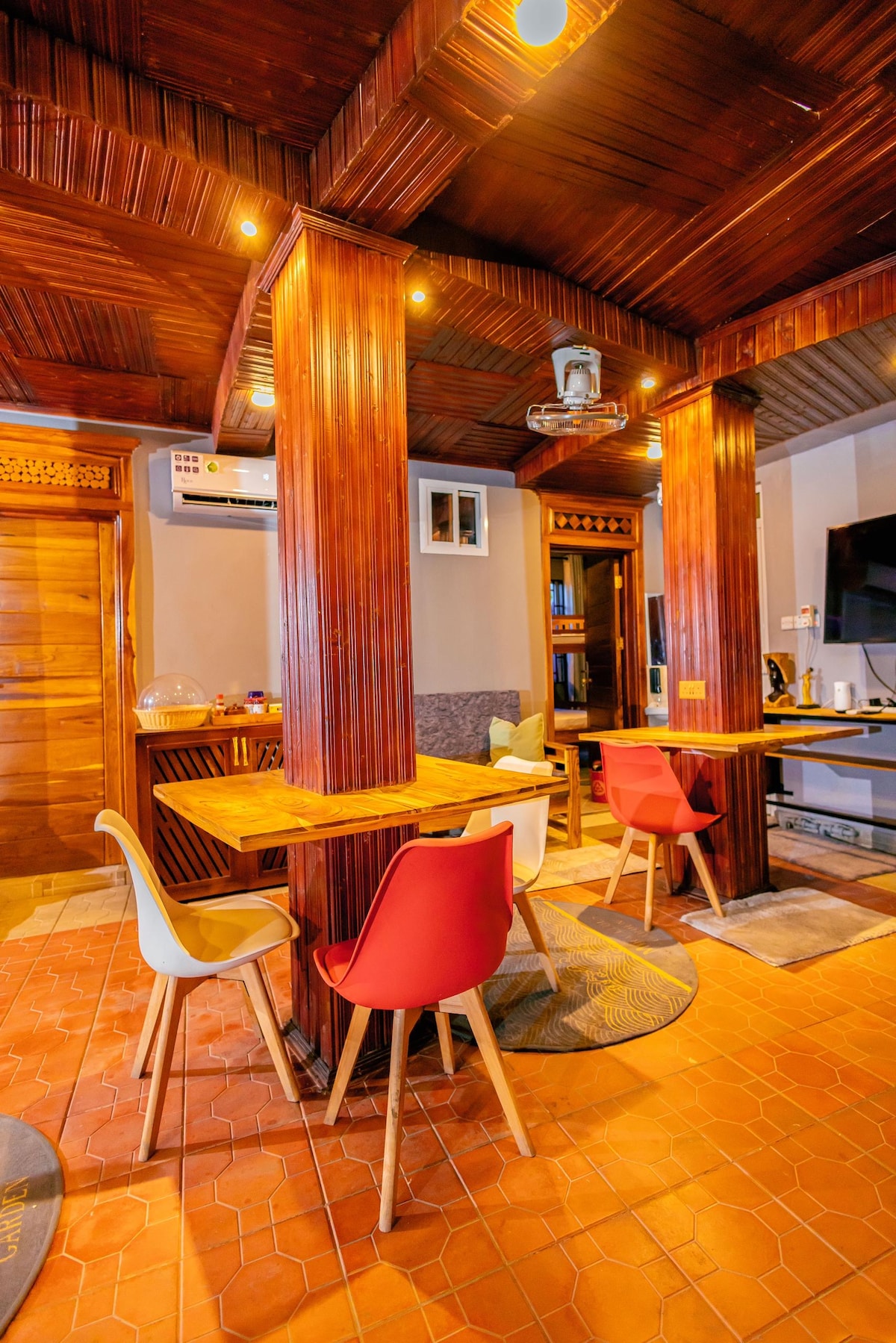 A dining area is showcased with a wooden table surrounded by four chairs in vibrant colors. Wooden pillars support a high ceiling, and warm lighting is present, creating an inviting space. A cozy living area can be glimpsed in the background, contributing to the overall ambiance.