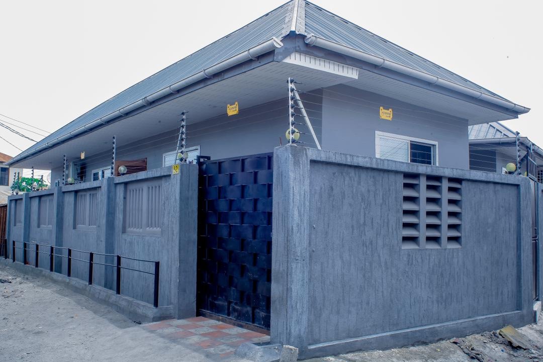 The exterior of the accommodation is characterized by a modern gray facade with a geometric brick entrance. A secure gate provides access, while low walls create a defined perimeter. Decorative lights are positioned along the top, enhancing visibility during the evening.