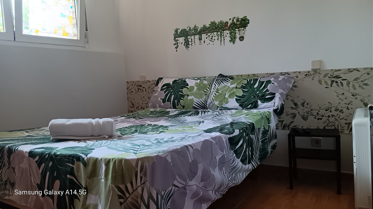 A spacious bed with a floral-patterned duvet cover is positioned against a wall adorned with greenery. A bedside table is located next to the bed, and natural light enters through a window, enhancing the bright and tranquil atmosphere of the room.