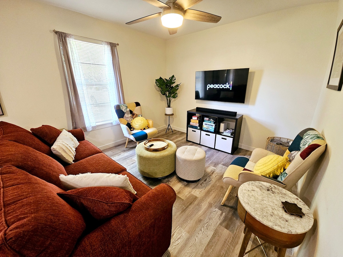 A welcoming living room is presented, featuring a spacious red sofa and a light-colored lounge chair with decorative cushions. A large TV is mounted on the wall, displaying the Peacock logo. A round ottoman and a small side table add functional charm, while natural light brightens the space.