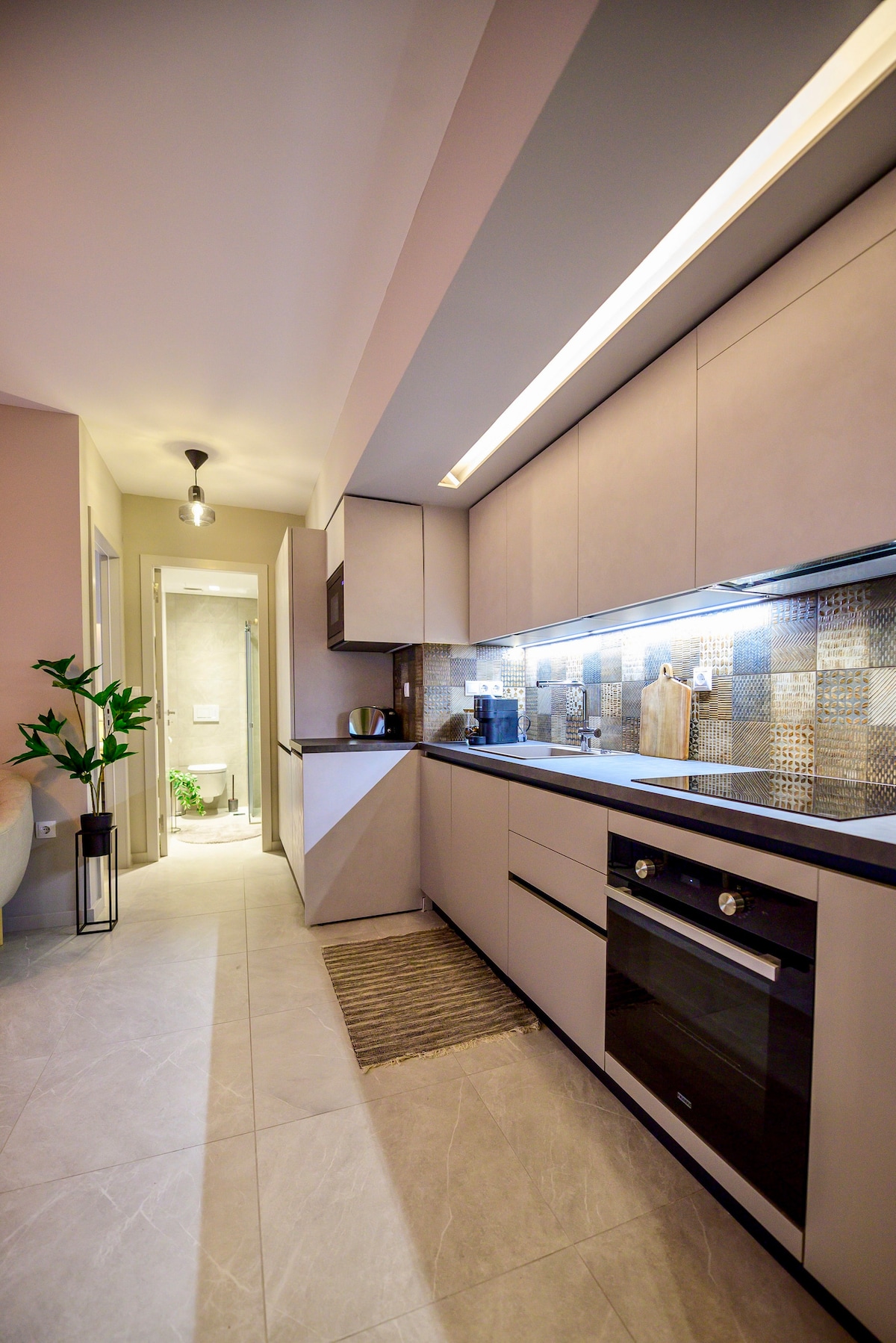 A modern kitchen is showcased, featuring sleek cabinetry and a spacious countertop. Equipped with an oven and a stylish backsplash of patterned tiles, the area is illuminated by under-cabinet lighting. Plants are placed to add a touch of greenery, enhancing the overall ambiance.