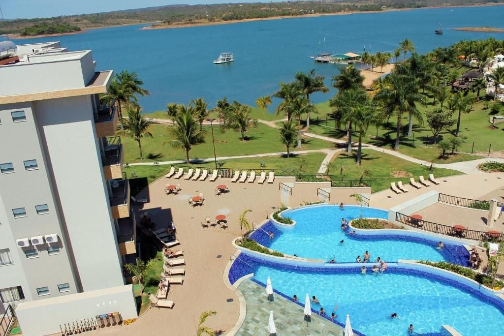 A spacious outdoor pool area is visible, with guests enjoying the water under clear skies. Sun loungers are arranged around the pool, while palm trees provide shade. A scenic lakefront is seen in the background, enhancing the serene environment.