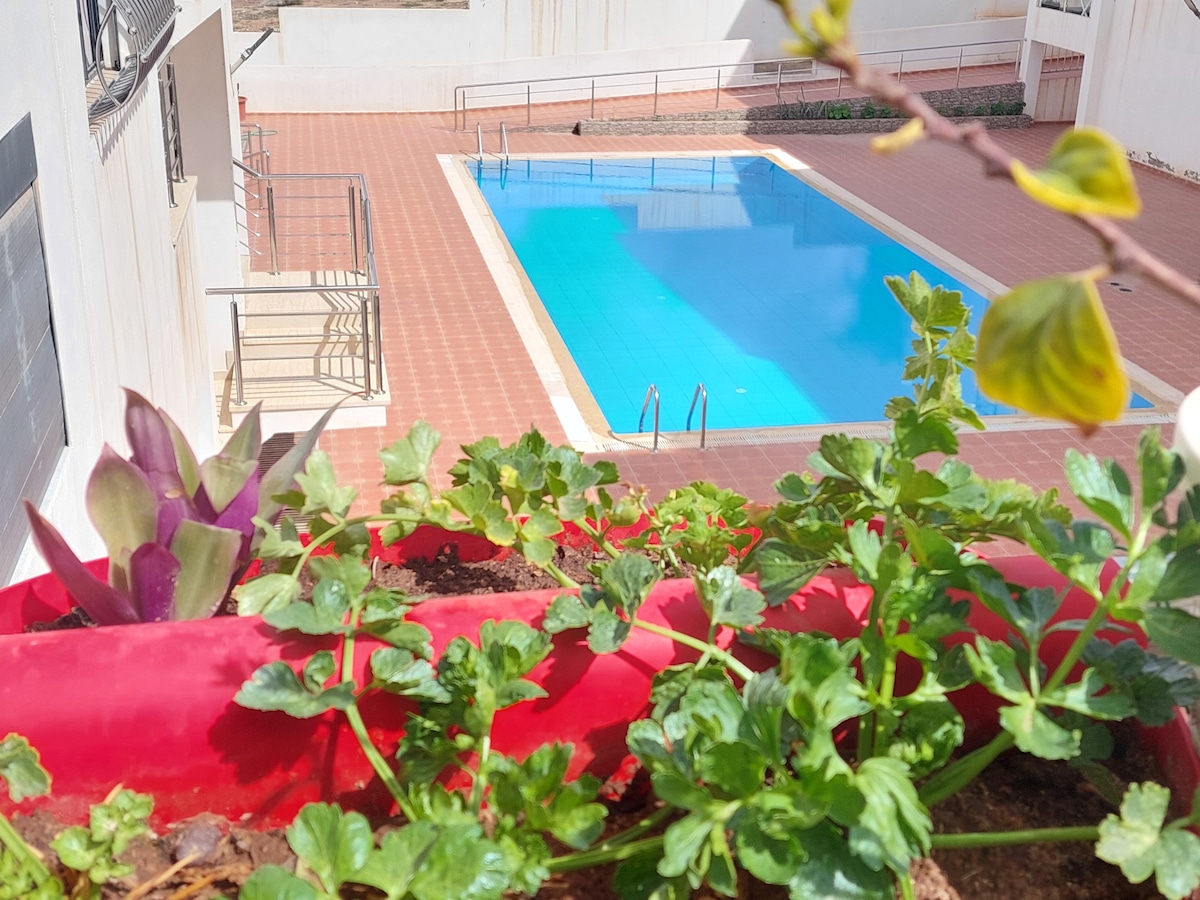 A clear blue pool is framed by a tiled deck and surrounded by a low wall. Lush green plants are visible in the foreground, adding a touch of nature to the scene. The area appears tranquil, ideal for relaxation.