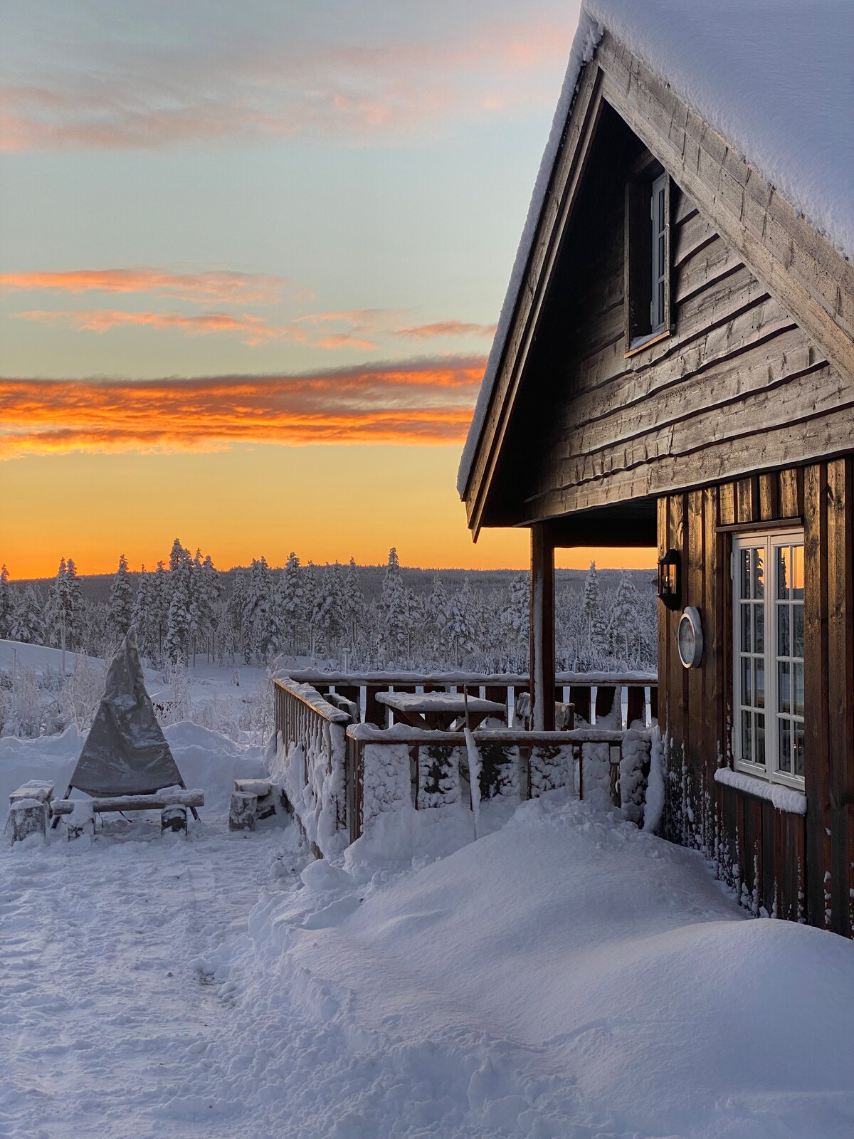 A wooden cabin is depicted with a sloped roof, adjacent to a deck overlooking a snowy landscape. A fire pit stands on the deck, framed by freshly fallen snow. The sky is illuminated with hues of orange and pink during sunset, enhancing the serene winter scene.