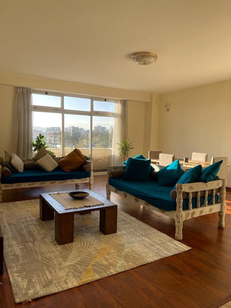A bright living area showcases a large window providing ample natural light. Comfortable teal sofas are arranged around a wooden coffee table, complemented by decorative cushions. A soft area rug anchors the seating, while a dining space with woven chairs is visible in the background.