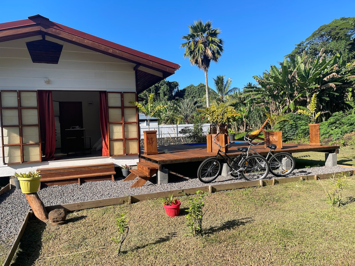 A charming bungalow is set amidst lush greenery, featuring a wooden deck with two bicycles resting against it. The entrance showcases a front porch with sliding doors, opening to the vibrant garden and tropical vegetation surrounding the property.