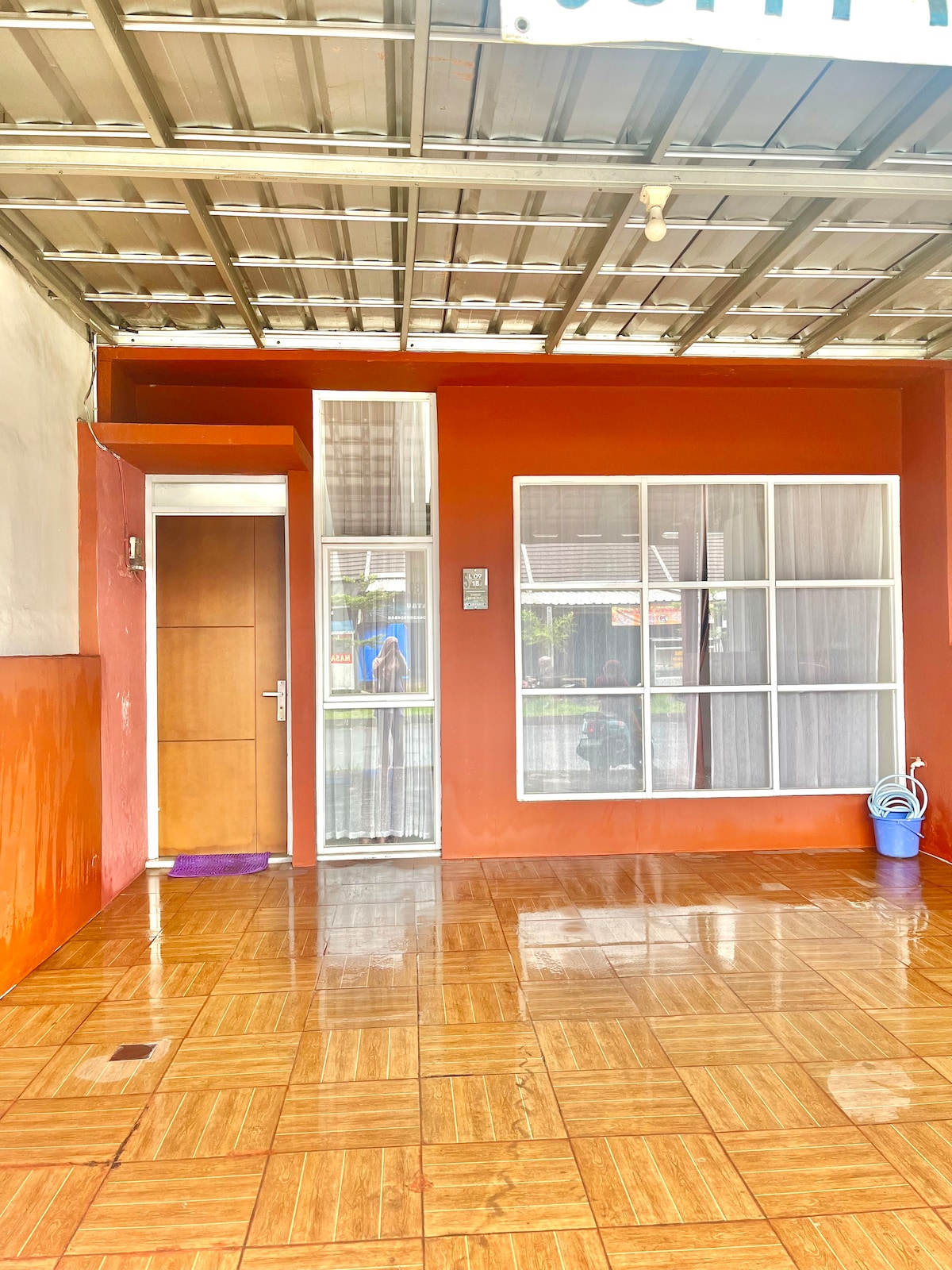 An entrance area features a well-maintained, glossy tile floor in warm wooden tones. A sleek wooden door with a glass panel leads to the interior. Large windows allow natural light to fill the space, complementing the orange-painted walls.