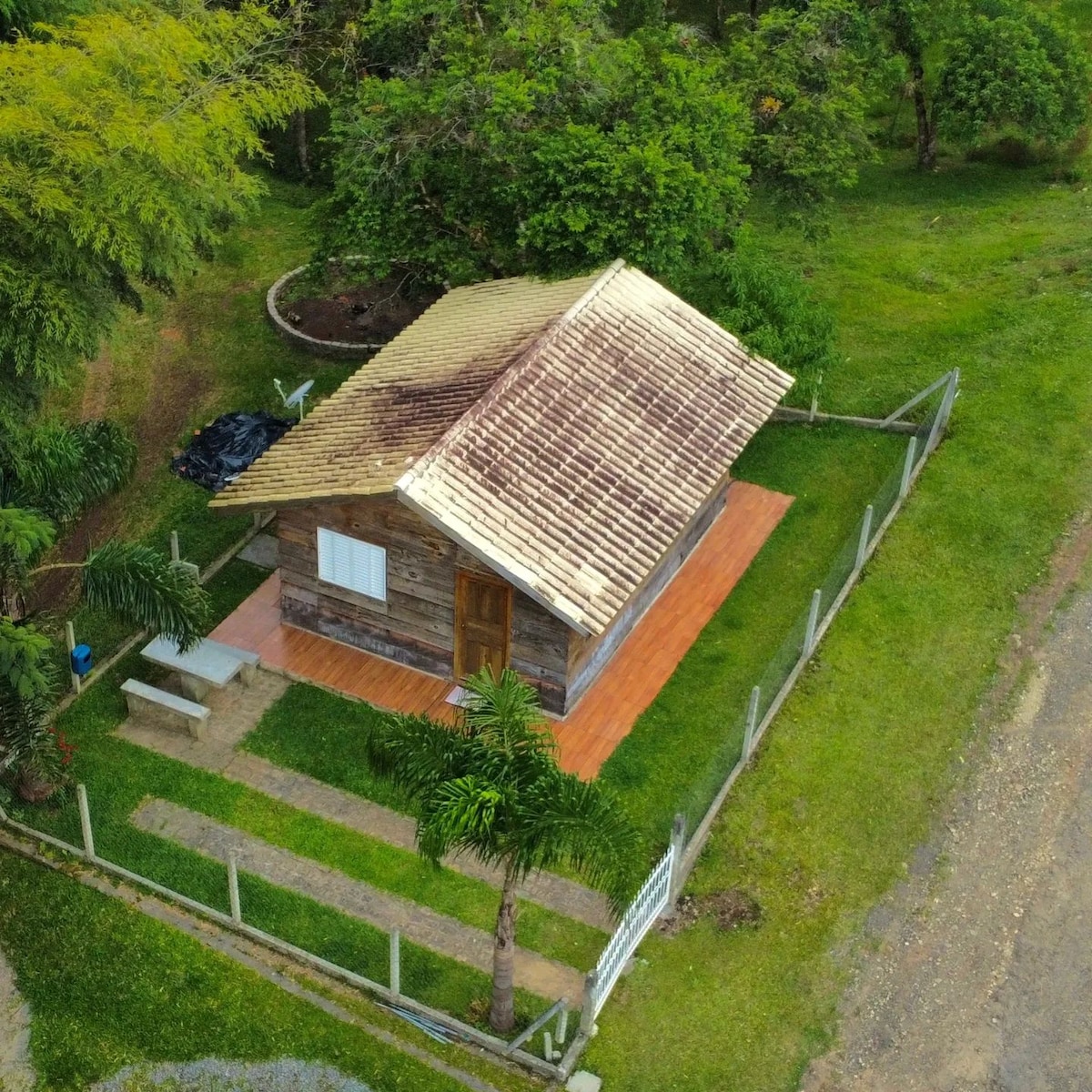 A rustic cabin is surrounded by greenery, with a sloped roof featuring wooden shingles. The cabin is positioned near a well-kept path leading to a patio area. A small outdoor seating arrangement is visible beside the entrance, promoting a peaceful outdoor experience.