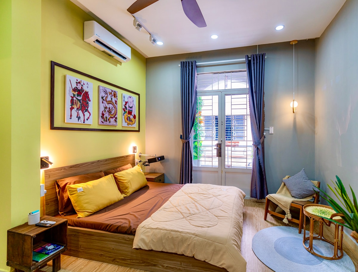 A cozy sleeping area features a spacious bed dressed with neutral-toned bedding, complemented by two decorative artworks on the wall. Natural light enters through a window, framed by dark curtains. A small seating area beside the bed includes a round table and a chair.