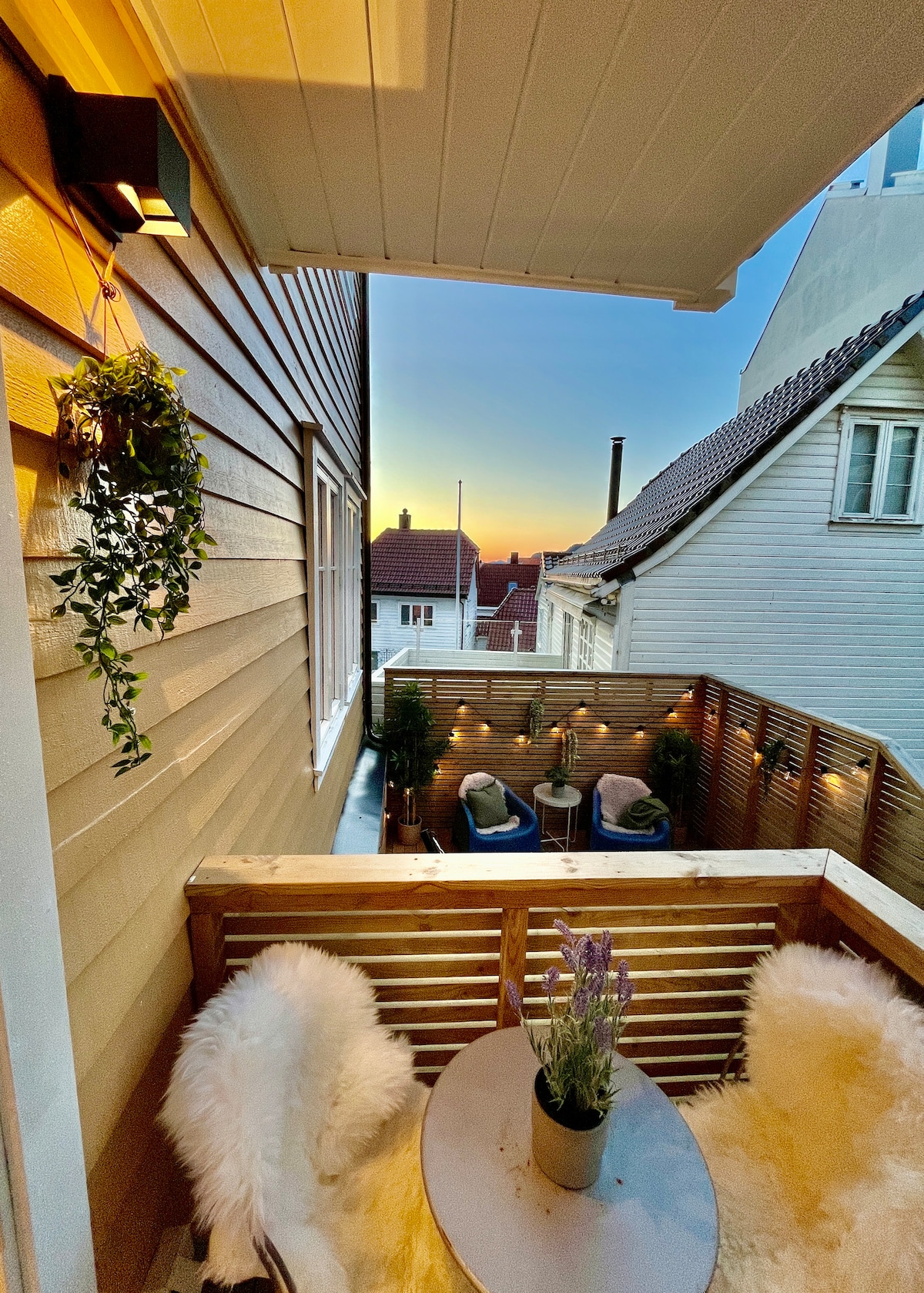 A private balcony is presented with wooden slats and adorned with potted plants. Two cushioned chairs surround a small round table, while soft lighting creates a calming ambiance as the sun sets in the background.