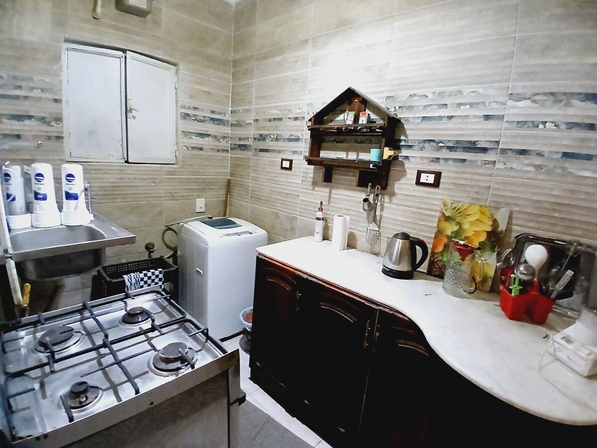 The kitchen features a gas stove and a stainless-steel sink with a counter space beside it. A washing machine is positioned against the wall, with shelves holding kitchen items above. Various appliances, including a kettle, are neatly arranged on the countertop.