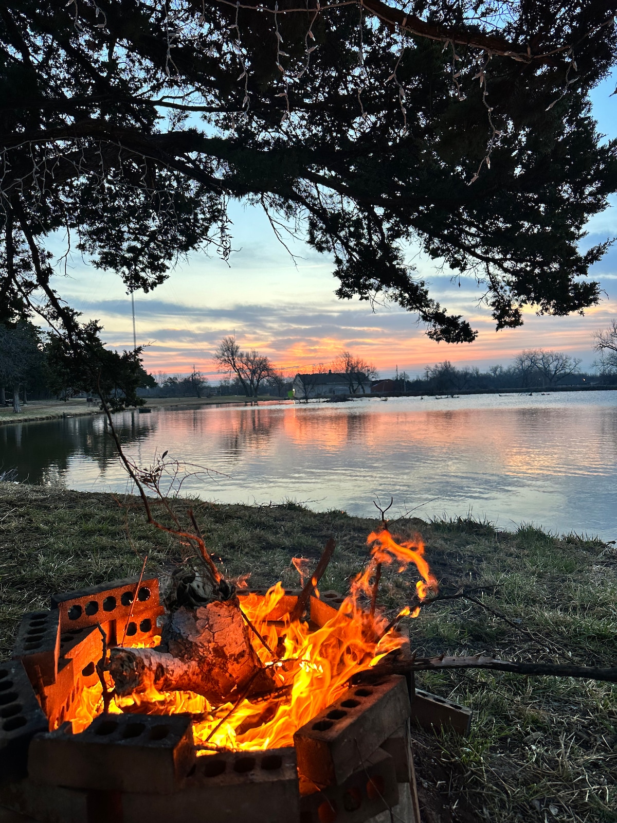 A warm fire burns brightly in a stone fire pit, surrounded by grass. The calm water reflects vibrant colors of a sunset, with trees framing the scene. Soft clouds drift across the sky, enhancing the serene atmosphere of the waterfront retreat.