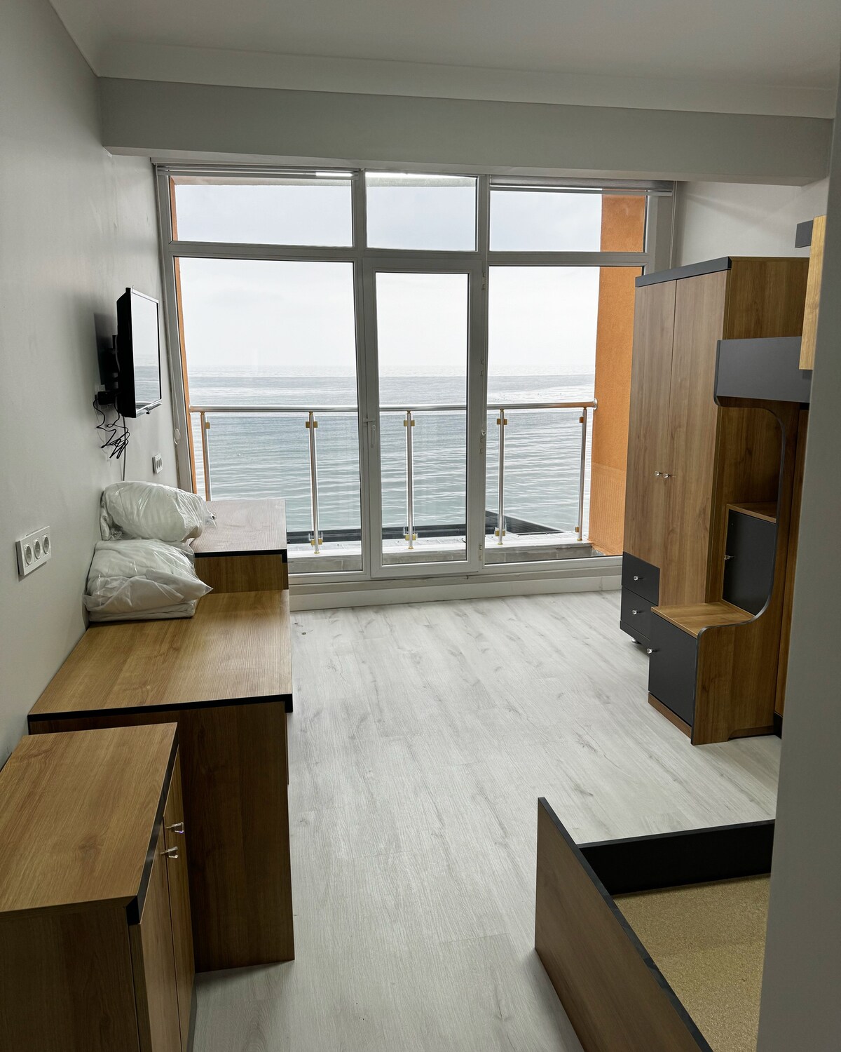 A bright room features large glass windows overlooking the water. Wooden furniture, including a dresser and storage units, is positioned strategically. Soft pillows are neatly placed on the bed, enhancing the room's functionality while allowing natural light to fill the space.