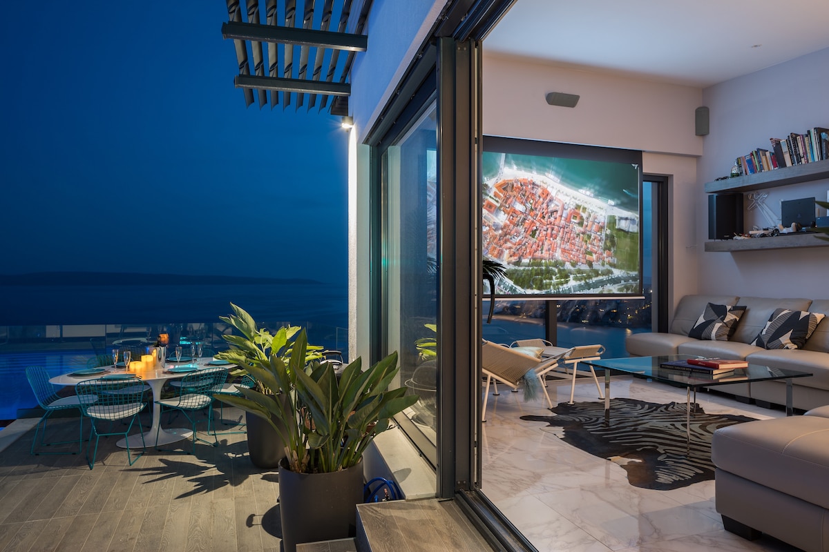 A modern open-plan living area features large glass doors that reveal an outdoor terrace. Lounge furniture is arranged for relaxation, and a sizable screen displays vibrant visuals. potted plants add a touch of greenery, complementing the sleek interior design.