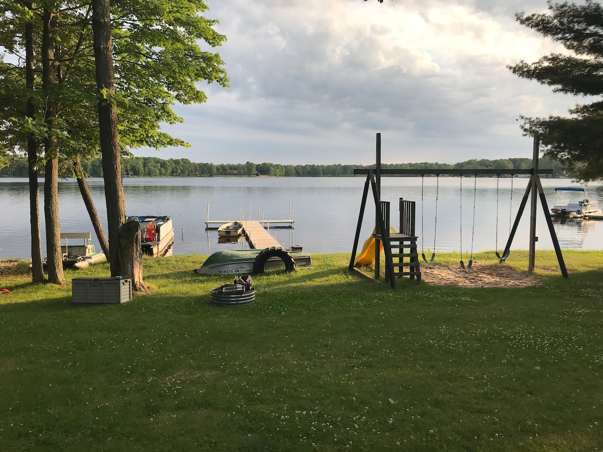 A serene lakeside view highlights the calm waters of Robinson Lake, with a dock extending into the lake. The grassy area features a playset, swings, and a small sandpit, surrounded by trees providing gentle shade.