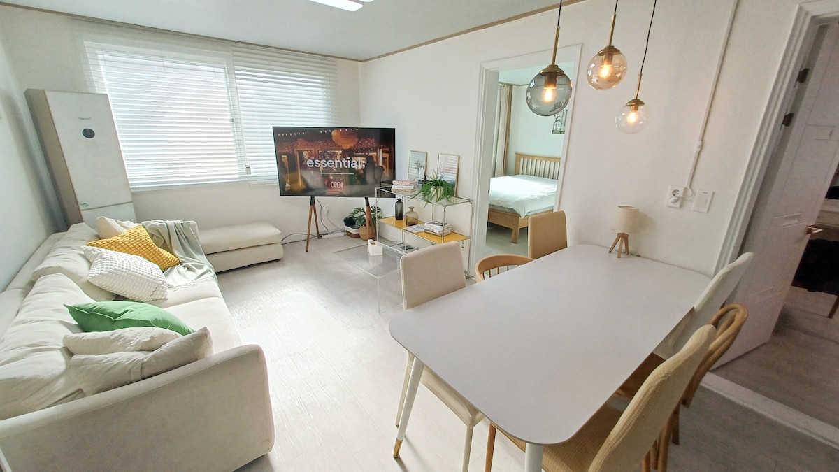 A spacious living area showcases a comfortable sofa adorned with cushions, facing a large TV. A modern dining table with wooden chairs is visible, and the room is illuminated by pendant lighting. Natural light enters through the window, enhancing the serene atmosphere.