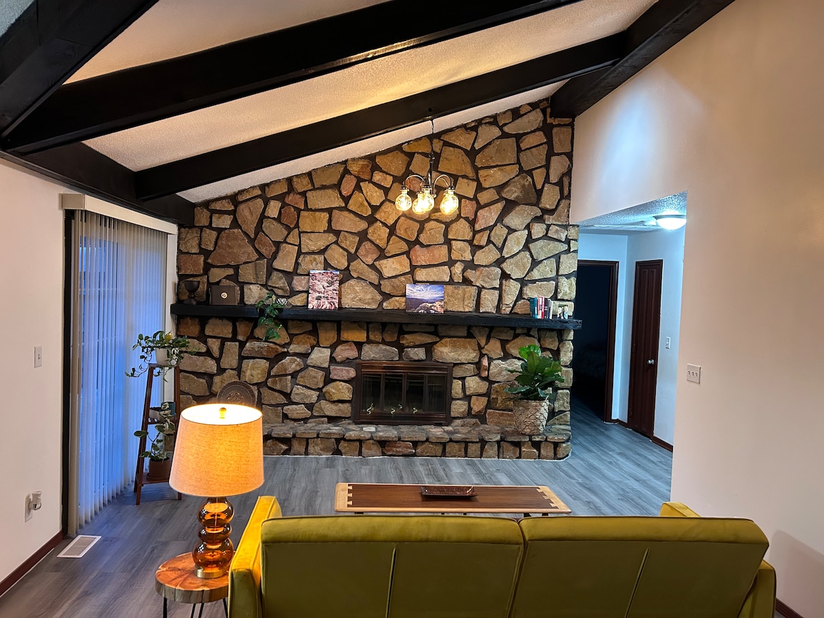 A spacious living area features a stone accent wall with a shelf for decor. A modern green couch offers comfortable seating under wooden beams. Soft light from a nearby lamp illuminates the room, creating a cozy setting. Large windows provide natural light and views of the outdoors.