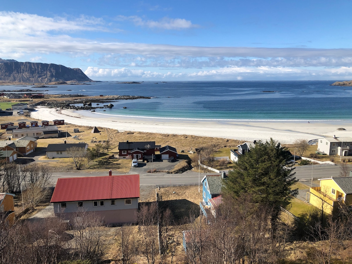 A panoramic view of the coastal landscape features a sweeping white sandy beach meeting a calm blue sea. Numerous houses, some with colorful roofs, are positioned along the shoreline, while distant mountains create a striking backdrop under a clear sky.
