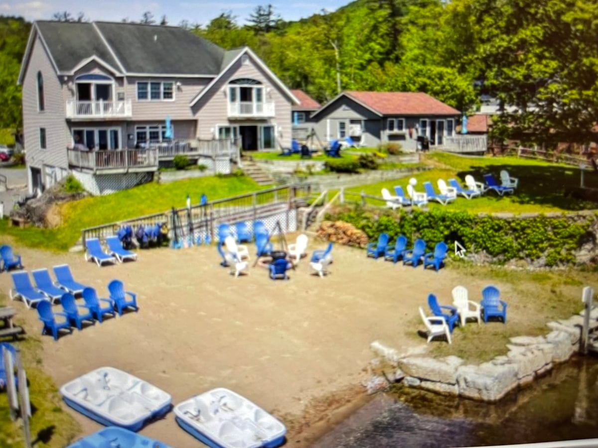 A sandy area features a variety of blue and white lounge chairs arranged for relaxation. Multiple paddle boats are docked near the water's edge, while a grassy space with trees provides a natural backdrop to the two-story cottage and neighboring building.